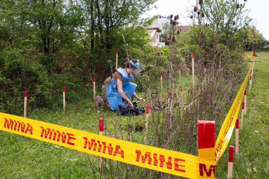 A woman wearing a large apron and face shield kneels down on grass, training to dig up explosives.