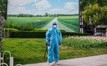 A security guard wearing a Hazmat suit stands in front of a patch of green and a large photo of rolling fields in Beijing