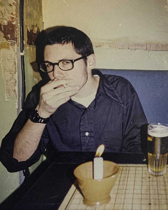 a color photograph of a man taking a drag of a cigarette and looking at the camera