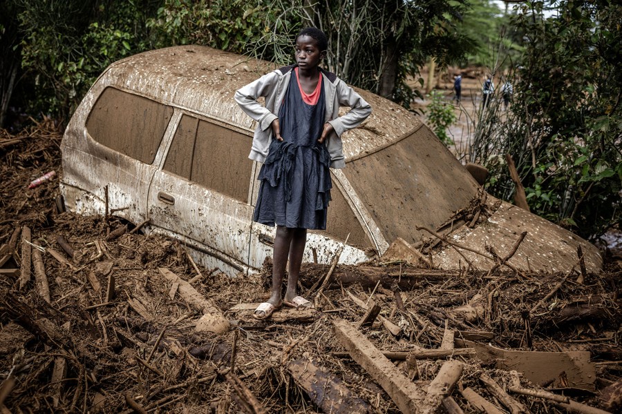 A girl stands, hands on hips, beside a wrecked car half-submerged in mud and dense flood debris.