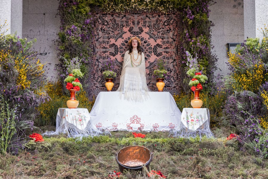 A girl sits on an altar, surrounded by flowers and plants.