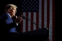 Donald Trump stands at a lectern in front of a flag. He is spotlighted; the rest of the scene is dark.