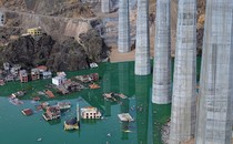 An aerial view of steep valley that is partially flooded, with several engulfed buildings and tall piers for a road bridge that is under construction.