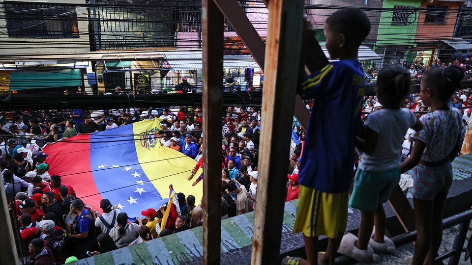 Photograph of three children on a balcony watching a march on the street below, with marchers holding a large Venezuelan flag