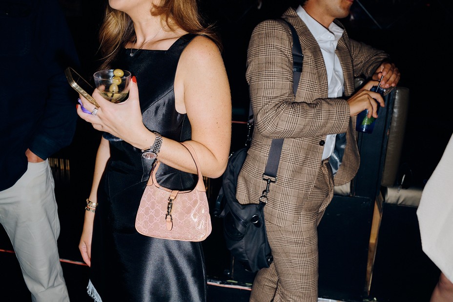 A color photograph of a woman in a black dress holding a martini and a man with his back to her in a plaid suit