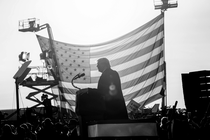 Fotografía en blanco y negro de Donald Trump dando un discurso ante una multitud y frente a una gran bandera estadounidense suspendida de dos elevadores de cangilones.