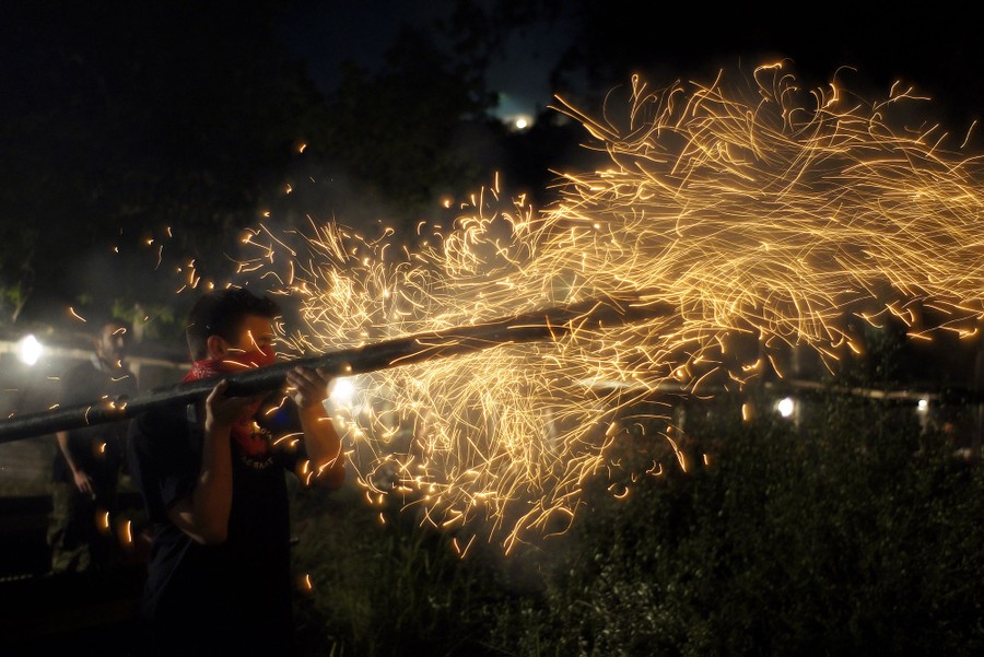 A person holds a long plank of some sort on their shoulder, using it to launch a rocket, which leaves many sparks in the air behind it.
