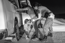 Una fotografía en blanco y negro de una mujer con una camiseta y jeans apoyada con ambas manos contra una camioneta blanca,mientras una mujer uniformada la inspecciona y una niña pequeña llora, por la noche.