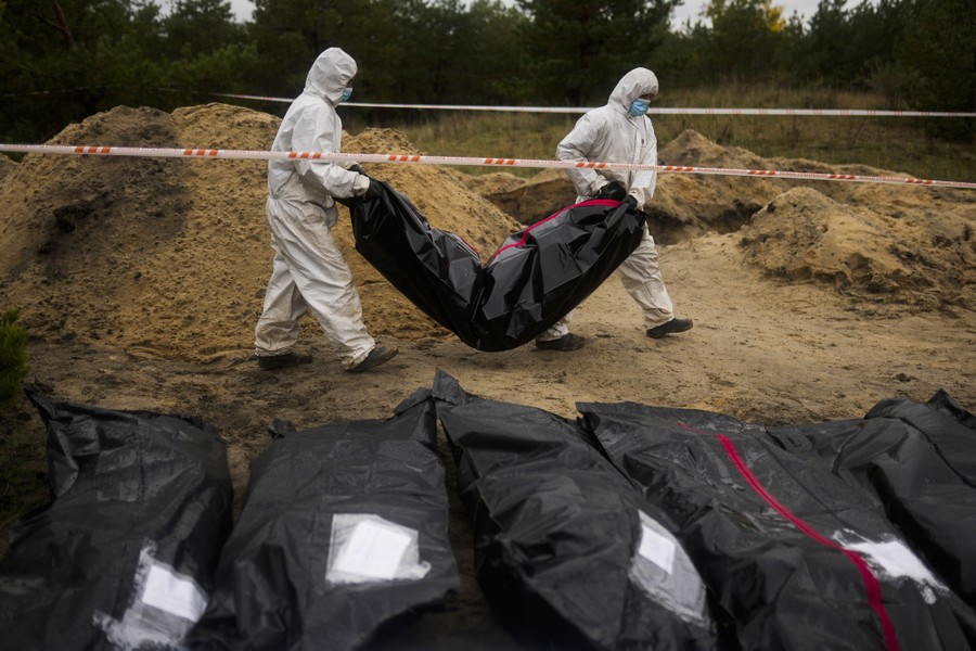 Two workers carry a body bag past piles of dirt and several other body bags lined up on the ground.