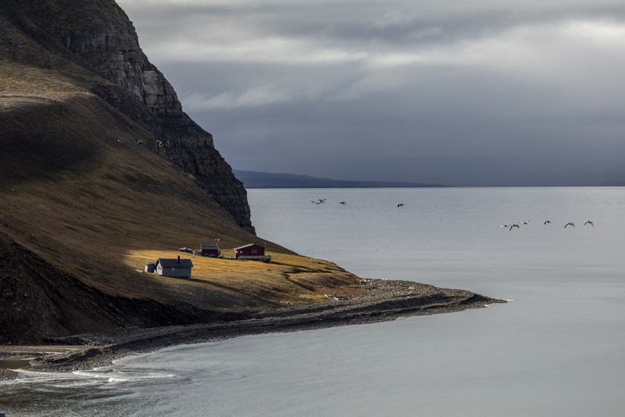 Houses stand at the foot of a steep mountain, along a shoreline.
