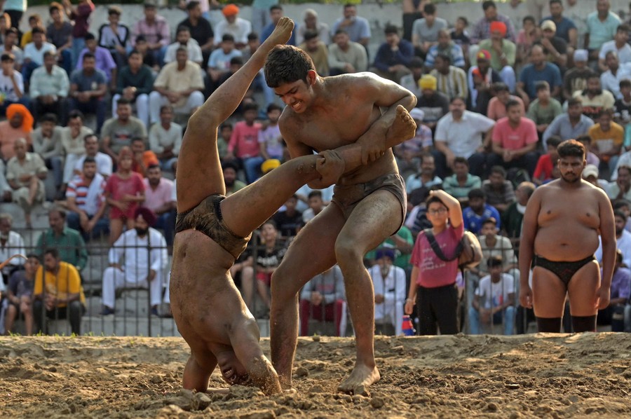 Two men wrestle in a muddy arena as a crowd watches from stands.