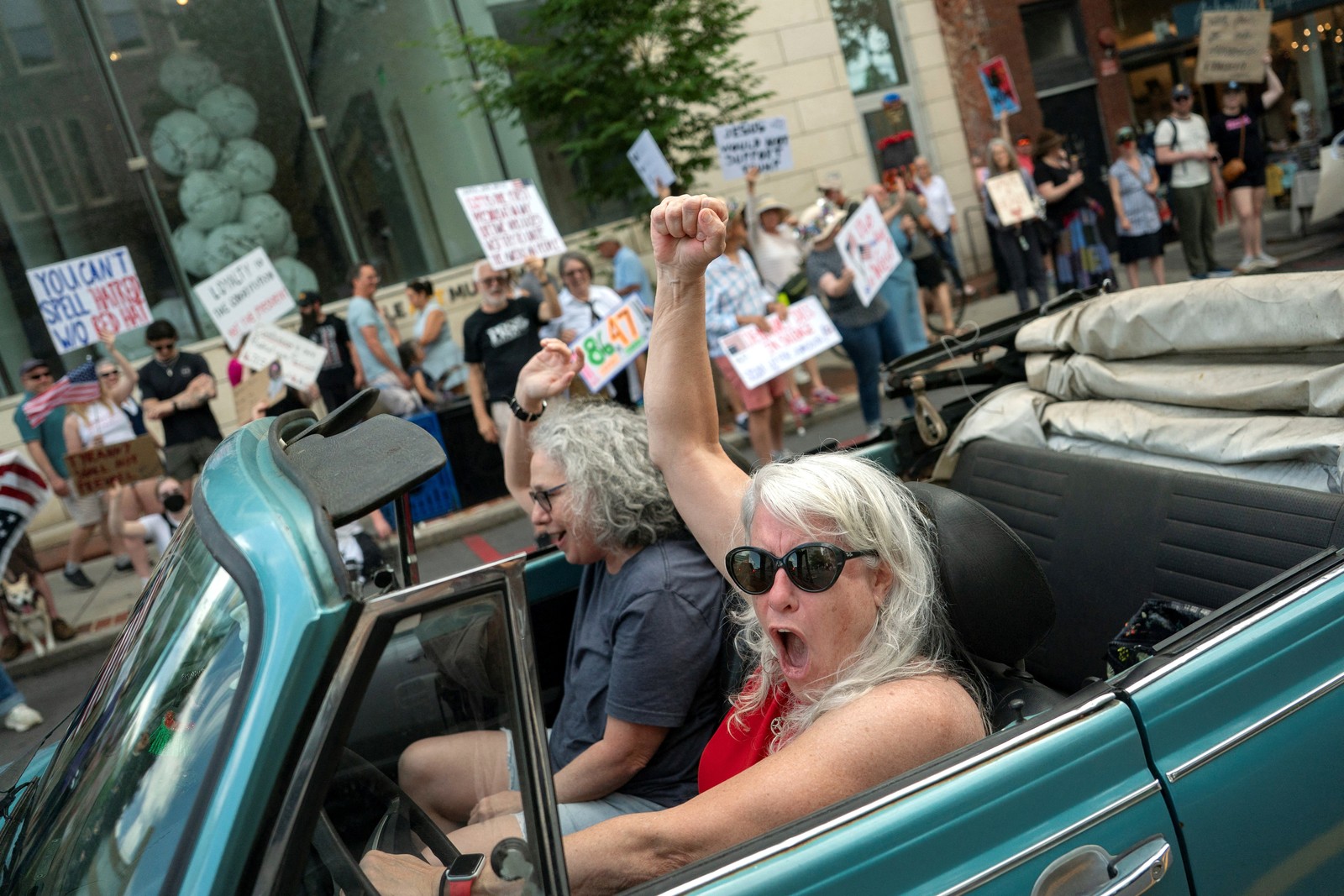 A driver in an open convertible car raises their arm, cheering, while passing a line of protesters.