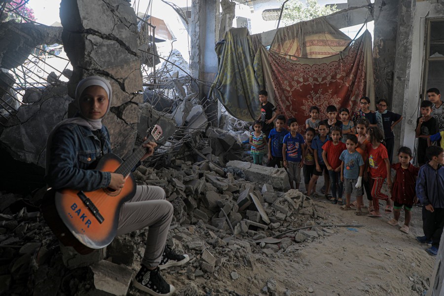 A young person sits on the rubble of a destroyed building, playing a guitar, as a crowd of children watches nearby.