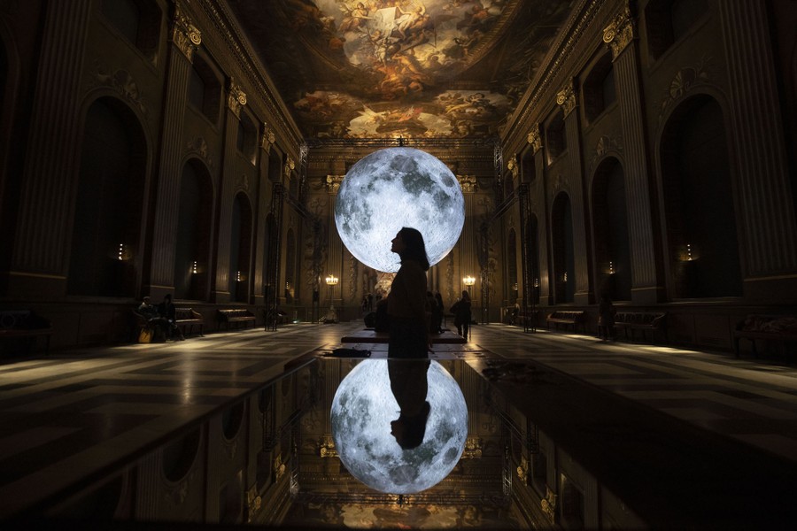 People walk beside a large illuminated moon hanging inside an ornate hall.