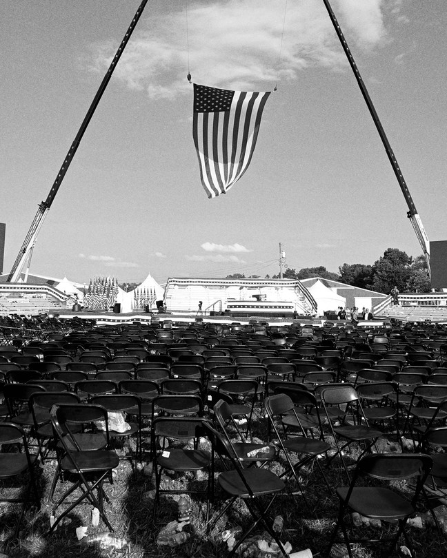 A black-and-white photograph of a large American flag being hung vertically over the stage of Donald Trump's Pennsylvania rally