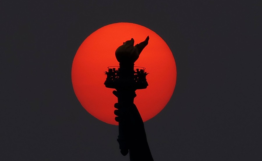 The torch in the hand of the Statue of Liberty is silhouetted by a reddish-colored sun.