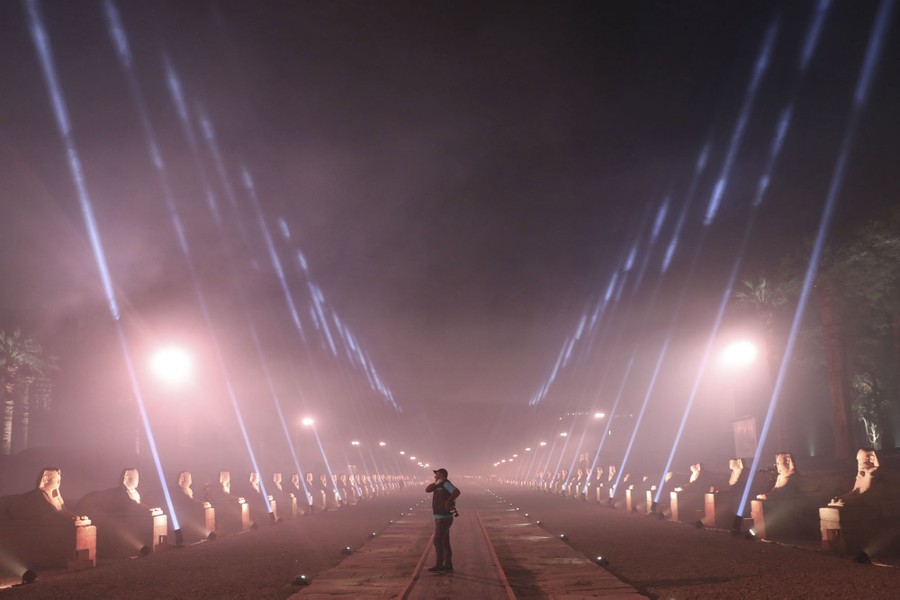 A person stands in the middle of a long avenue that is lined with sphinx statues and spotlights.
