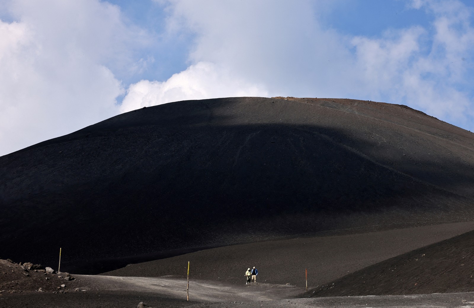 Two tourists walk in a vast hilly area covered with dark volcanic rock and ash.