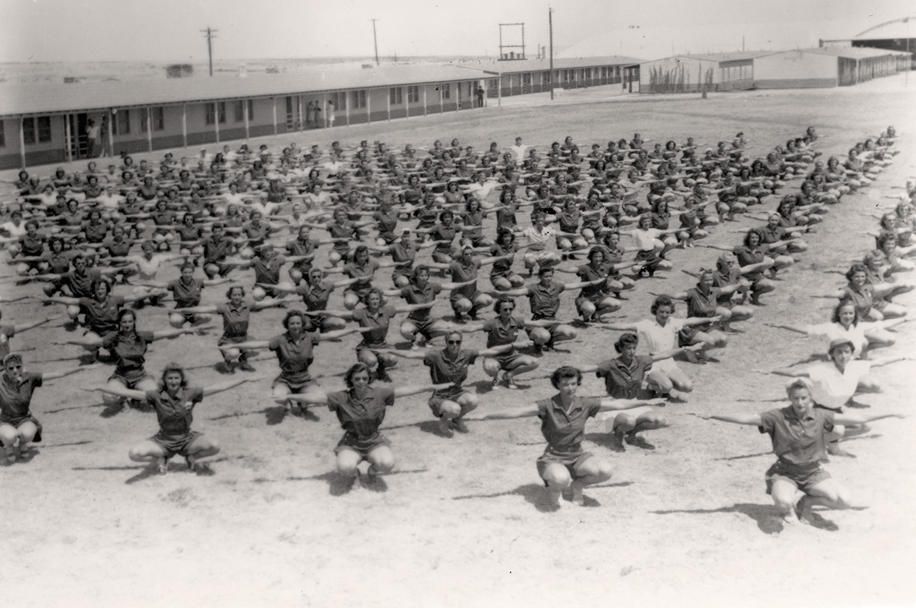black-and-white archival photo of dozens of women exercising in rows on field, squatting with arms stretched out 