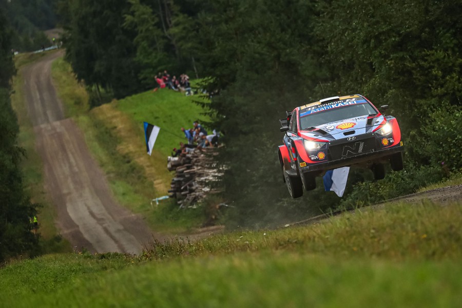 People beside a dirt road watch as a rally car passes at high speed, catching air.