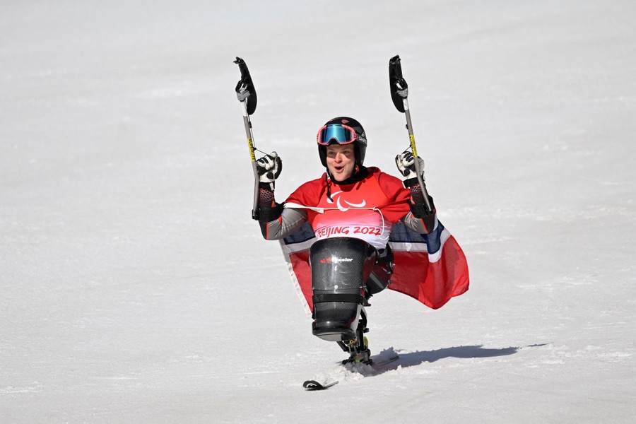 An athlete in a sit ski holds up his arms (holding outriggers) while wearing a Norwegian flag across his shoulders.