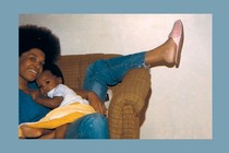 A woman sitting in a brown chair with her leg thrown over the arm holds a baby while looking at the camera.