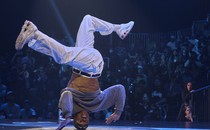 A dancer performs an acrobatic move in front of a crowd, while upside down, supported by only their head.