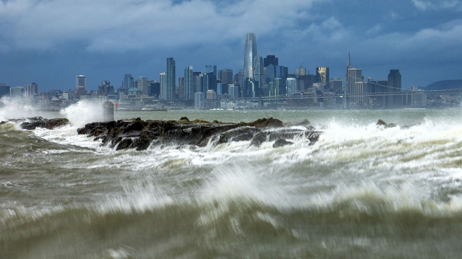 Waves crash over a breakwater, with the San Francisco skyline as a backdrop.