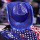 A color photograph of a person in a sparkly blue cowboy hat with an American flag draped around their shoulders, seen from the back.