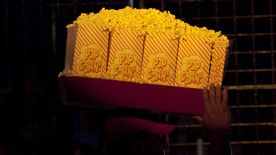 A vendor carries large bags of popcorn on a tray above his head.