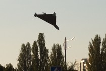 A large broad-winged drone flies towards buildings in a city, angled downward.