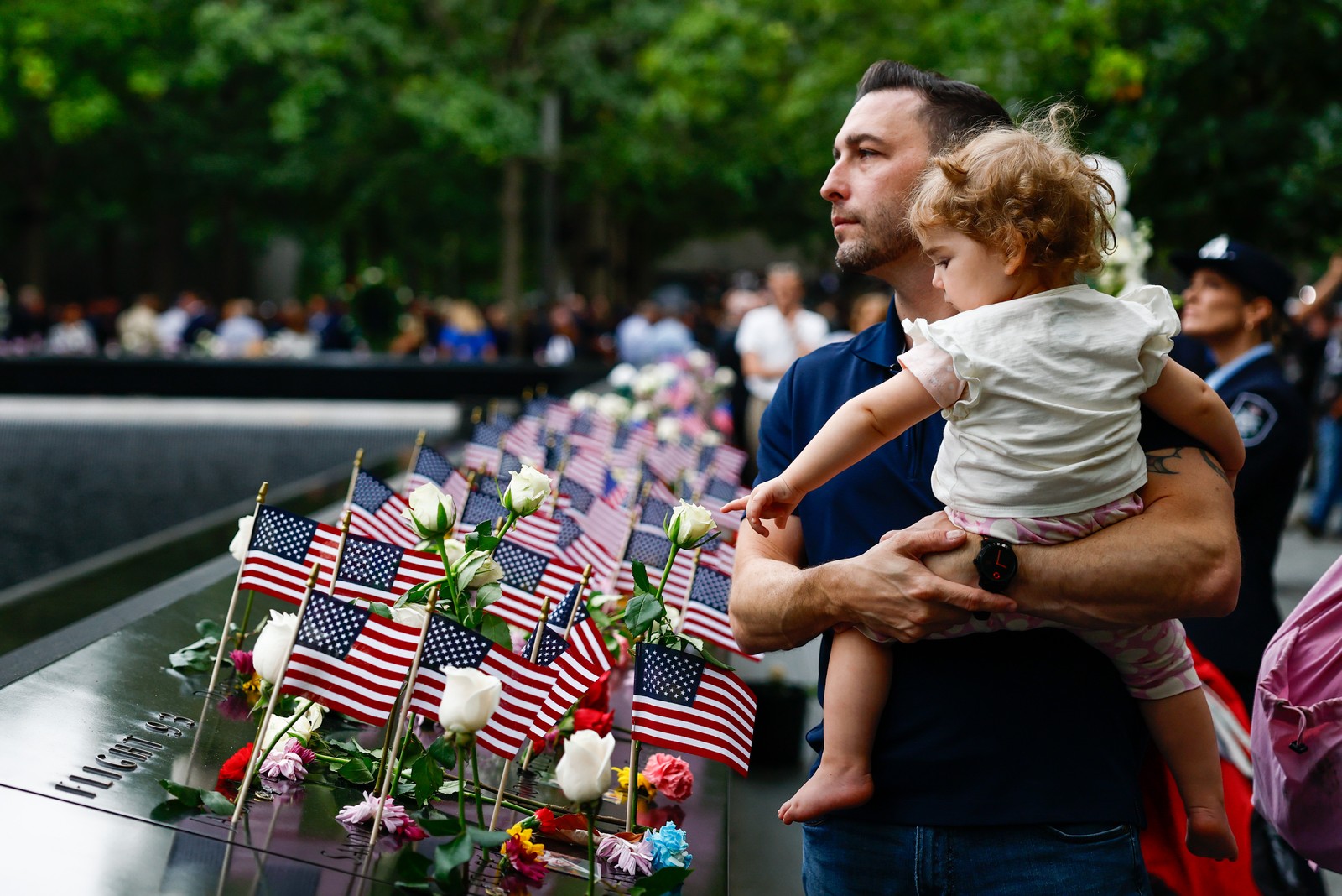 A man holds his young daughter, standing beside a memorial adorned with flowers and American flags.
