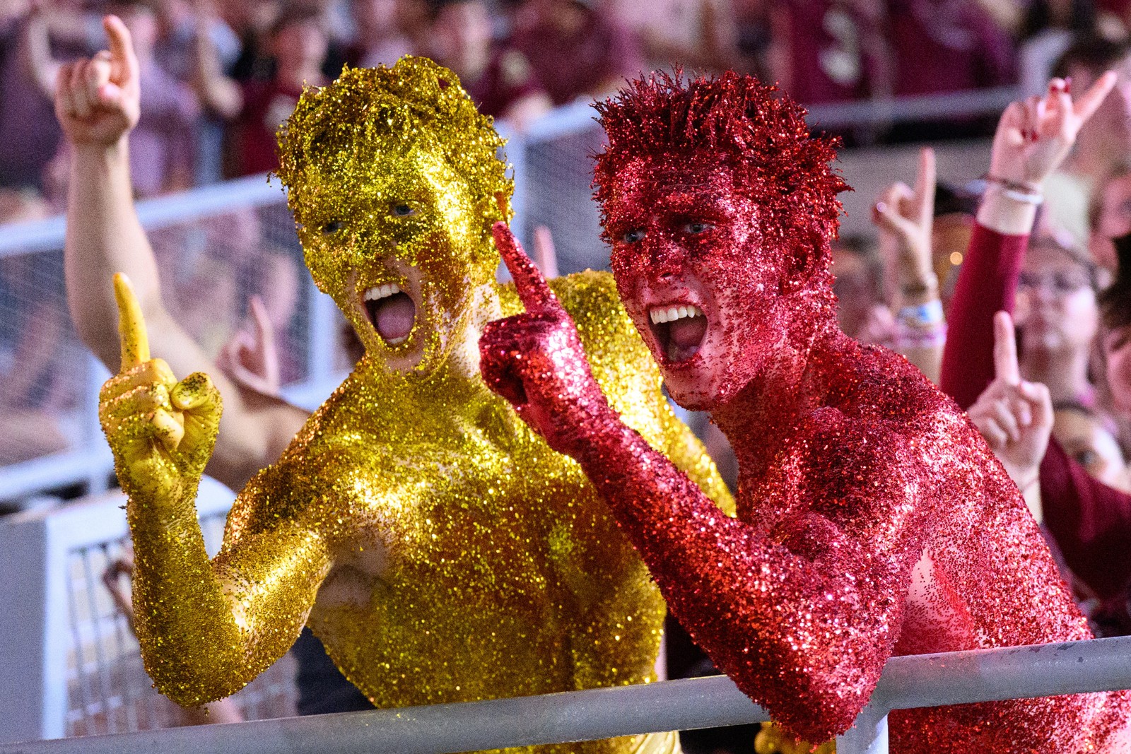 Two football fans, completely covered in colored glitter, cheer in the stands.