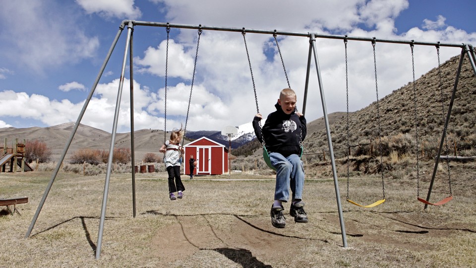 Two children play on the swings outside a red schoolhouse. The setting is rural.