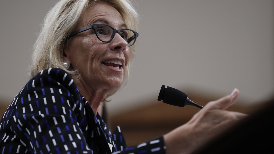 Betsy DeVos gestures while sitting at a microphone