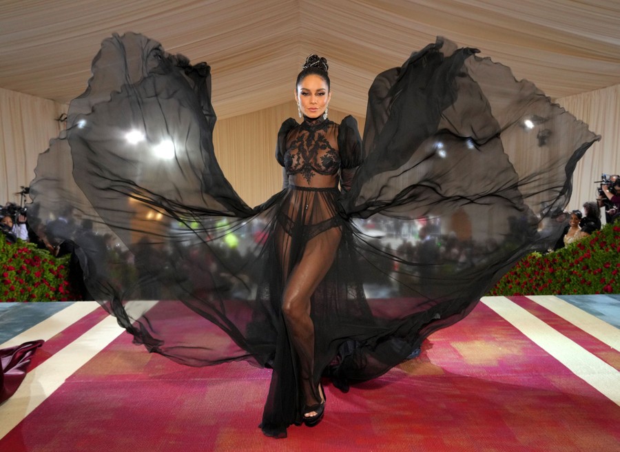 A woman wearing a sheer, flowing garment poses for photographers on a red carpet.