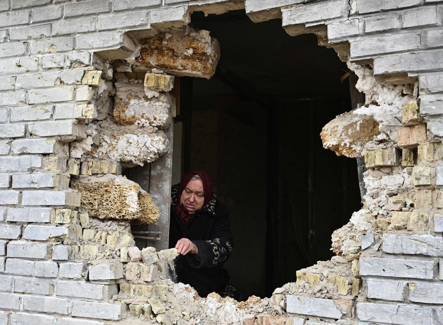 A person stands inside a damaged house, seen through a huge hole blown in a brick wall.