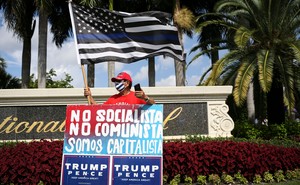 A man in a MAGA hat stands with a sign that reads "Not socialist, not communist, we are capitalist" in Spanish