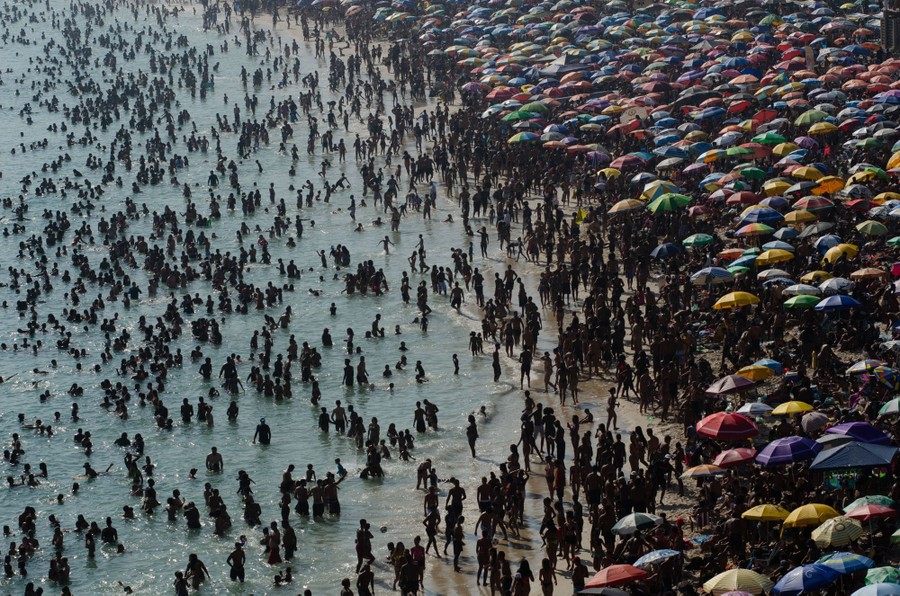 Thousands of people crowd along a beach, some in the water, others on the sand, under umbrellas.