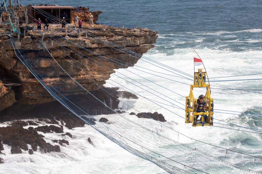 People ride in a small gondola that travels along cables strung between rocky cliffs, above crashing sea waves.