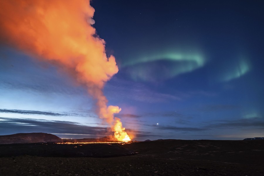A glowing lava flow illuminates a plume of gas and steam, with the northern lights above.