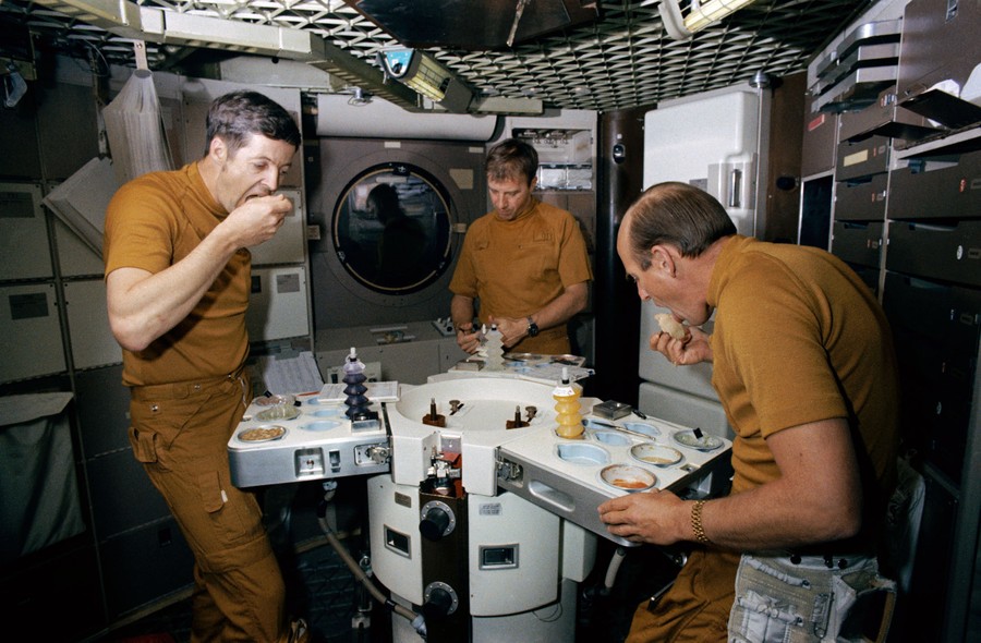 Three men stand inside a training mock-up of a space station eating food.
