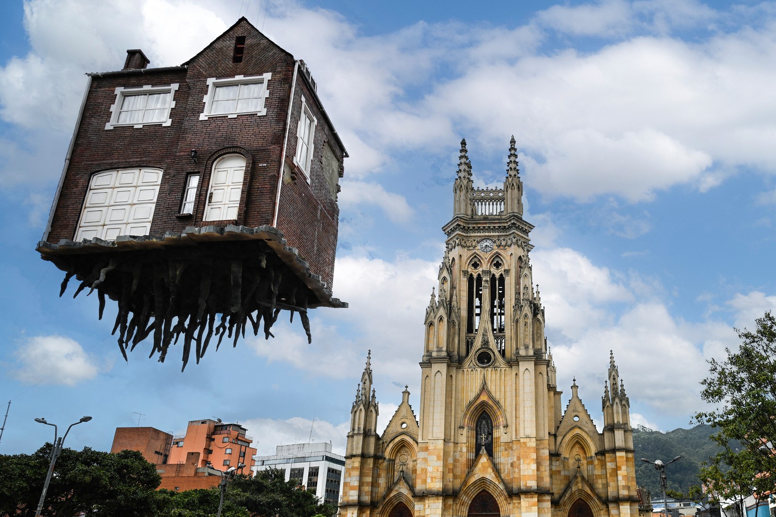 A large art installation, looking like a small, two-story brick dwelling that appears to be floating about 50 feet above a city square, with thick roots hanging beneath the structure