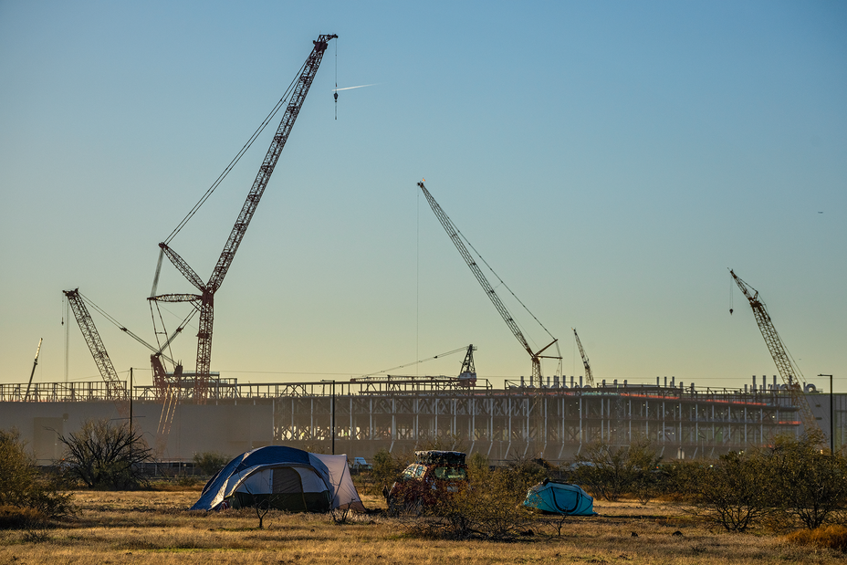 photo of massive construction project with multiple large cranes in background, with tents and desert scrubland in foreground