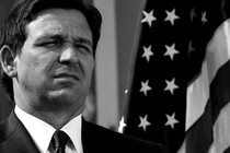 Black-and-white photo of Florida Governor Ron DeSantis squinting, standing in front of an American flag
