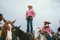 photo of Black boy in boots, jeans, pink long-sleeve button-down shirt, and cowboy hat, hands on hips, standing on the saddle of horse next to several other people on horseback