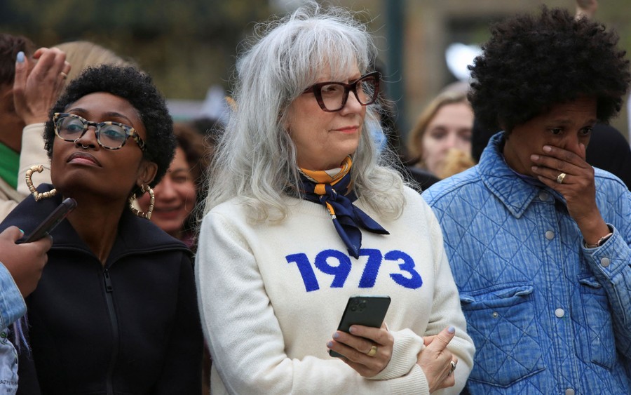 Three women standing among others during a rally. The one at center wears a sweater with "1973" written on it.