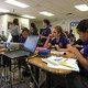 Students dressed in purple polos sit in a classroom with papers, pencils, and a laptop strewn in front of them.