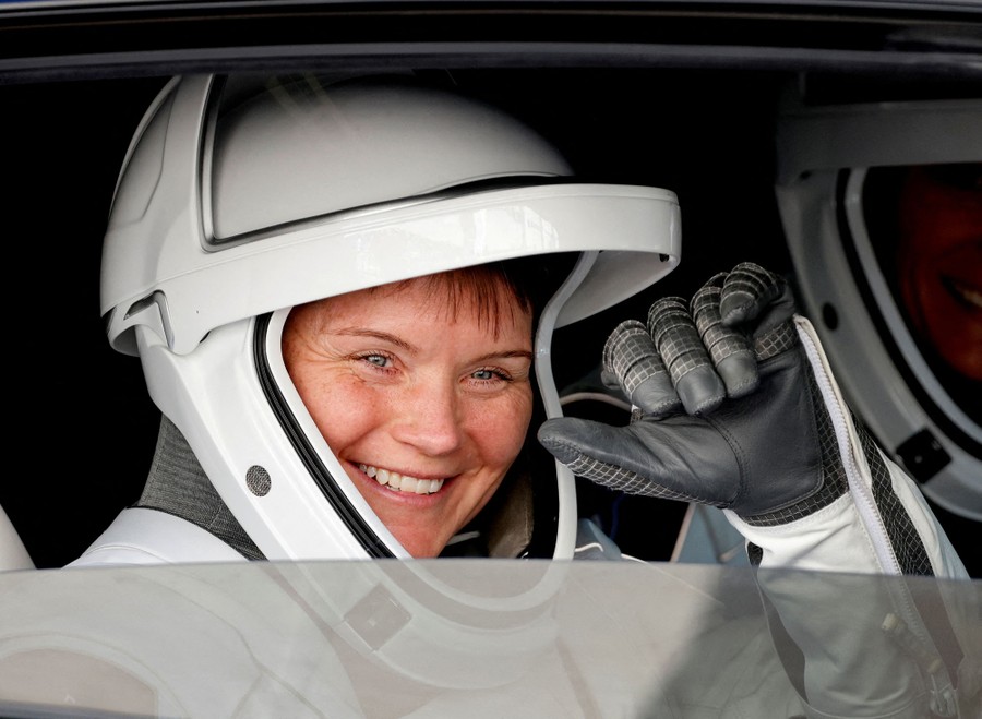 An astronaut wearing a large white helmet waves from inside a vehicle.