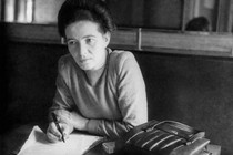 Simone de Beauvoir sits at a table at the Café de Flore, looking into the distance. She has pieces of paper in front of her and holds a pen. Her purse is on the table beside her.